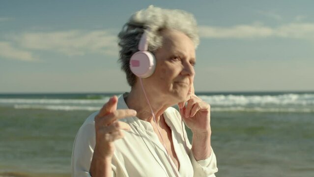 cinematic video of an elderly woman dancing on the beach near the water while wearing headphones and playing music.