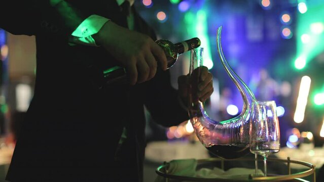 Crop of male person in formal outfit holding bottle of red wine and pouring drink in elegant glassy decanter on wedding banquet. Competent sommelier preparing alcoholic beverages for guests.