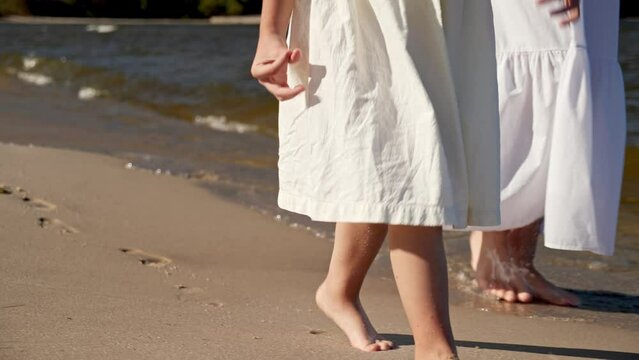 Mom and daughter in dresses walking along the beach, close-up of legs