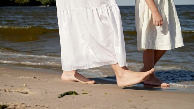 Mom and daughter in dresses walking along the beach, close-up of legs