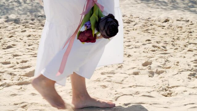 Legs of girl walks barefoot along the beach in a white dress with a bouquet of flowers