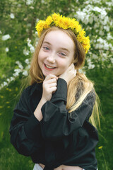 A girl of 13 with a wreath of dandelions in a spring flowering garden. The girl smiles and looks at...