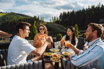 Group of friends drinking cocktails and enjoying meal while sitting on restaurant terrace in mountains - obrazy, fototapety, plakaty