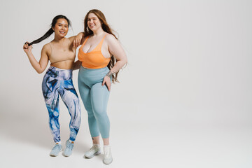 Two cheerful women in sportswear hugging each other while standing isolated over white wall