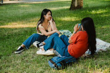 Asian woman taking picture of her friend with instax camera while sitting on grass in park