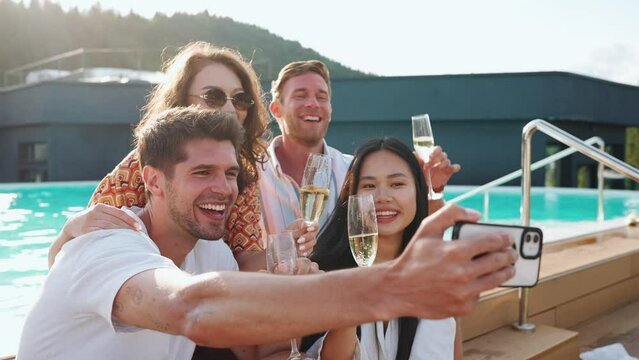Smiling multinational friends taking selfies with phone and clinking champagne glasses by pool at mountain resort on balcony in summertime