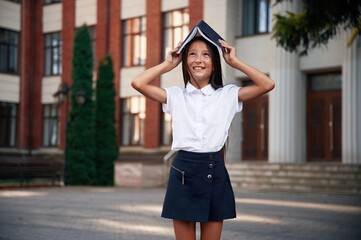 Playing with book that is over the head. School girl in uniform is outdoors near the building