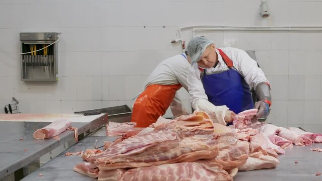 Butchers men cutting pork meat from bones for preparing delicacies in meat shop. Selected meat products in butchery, meat factory. Food plant, preparation raw production. Pork falling on metal table.