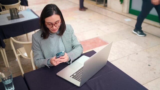 Businesswoman in jacket and glasses sitting in outdoor cafe and working on laptop and smartphone. Woman typing message on her cell phone. Online communication in street cafe. High angle