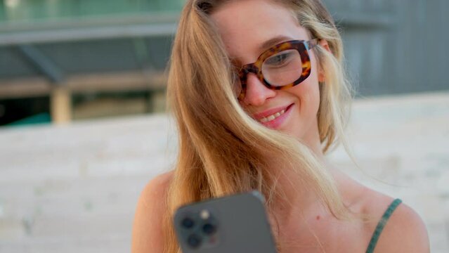 Pretty young blonde woman with glasses video chatting on smartphone sitting on stairs outdoor. Happy girl talking to his friends on social networks during free time. Online communication
