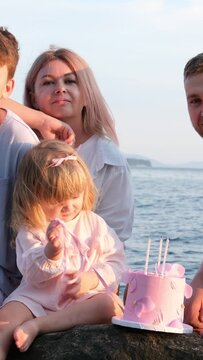 celebrating the birthday of small child on ocean shore girl playing knocking on cake parents trying to focus on camera look at frame smiling girl is busy with own business. hitting candles with ribbon
