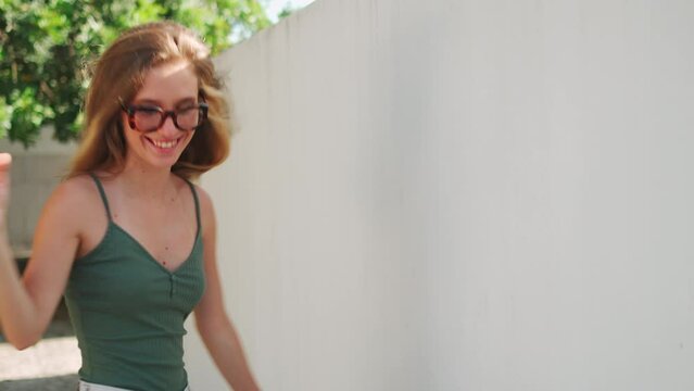 Back view young blonde caucasian woman walking down street by white wall and guiding her hand along it. Attractive blonde girl in glasses turning to camera with smile. Summer walk around city
