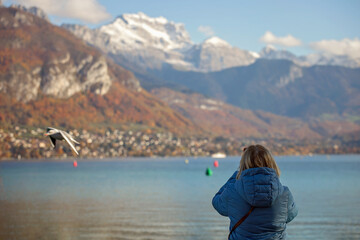 Fototapeta na wymiar A senior woman stands by Lake Annecy, capturing the mountainous landscape on her mobile phone. Elder traveler in the serene beauty of the French Alps