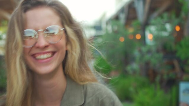 Back view of young caucasian blonde girl walking cafe outdoor terrace and turning to camera with smile. Happy young woman in glasses on greenery and street garlands background on summer evening