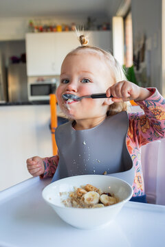 Happy little girl in high chair feeding herself breakfast with a spoon