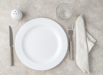 White empty ceramic plate with fork and knife, glass, salt and napkin over stone background