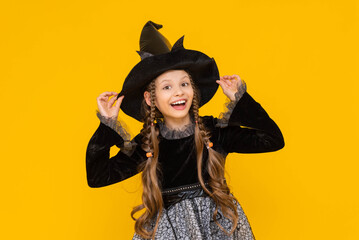 A funny and joyful child in a Halloween costume, on an isolated yellow background. A happy little girl in a witch costume and a witch hat is having fun at a Halloween party.