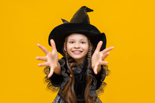 The little sorceress pulls the handles forward and scares. A witch in a carnival costume for Halloween, a beautiful young girl in a lush black dress for a party, a witch on an isolated background.