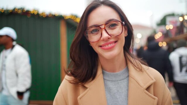 Portrait shoot smiling female tourist in beige coat and glasses european Christmas market on warm winter day. Elegant young woman fixing his eyeglasses and looking camera. Happy people 