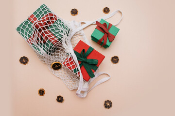 Red and green gift boxes lie in an eco-friendly white string bag on a beige background with dried manadrins. view from above. Space for copying
