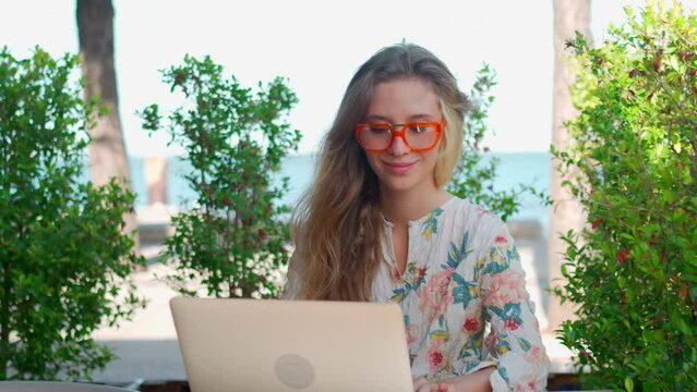 Attractive businesswoman sitting with laptop on terrace with sea view on sunny summer day. Modern female freelancer in glasses and beautiful dress enjoying breeze. Remote work in pleasant atmosphere