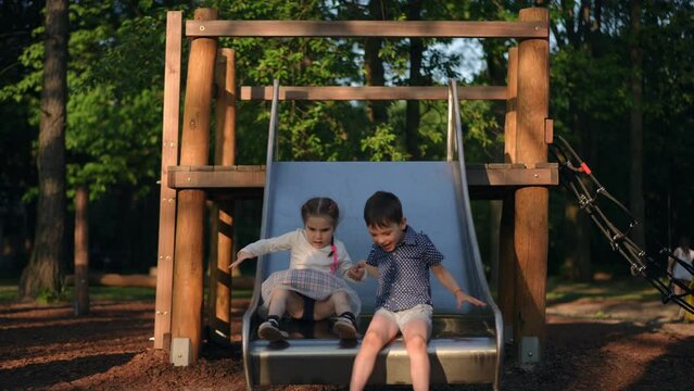 Boy and girl sliding down slide on playground in slow motion holding hands. Front view portrait of relaxed Caucasian happy friends enjoying leisure having fun playing outdoors at sunset in park