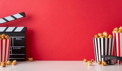 Cinematic anticipation: A side view table displays striped boxes filled with cheese and caramel popcorn, 3D glasses, and a clapperboard against a red wall, setting scene for a movie premiere gathering