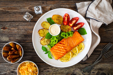 Seared salmon steak with baked potatoes and fresh vegetable salad served on wooden table
