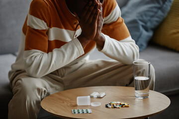 Closeup of depressed Black man sitting on couch at home with pile of prescription pills on table, copy space
