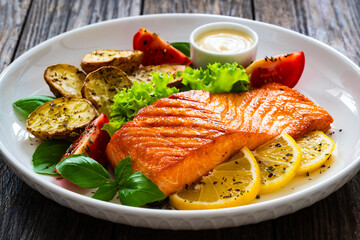 Seared salmon steak with baked potatoes and fresh vegetable salad served on wooden table
