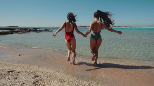 Two fit beautiful young mix race asian women in red swimsuits run into water at the beach blue lagoon sea coastline. Splashing water in slow motion. Girls tourists spend time together. Luxury resort.