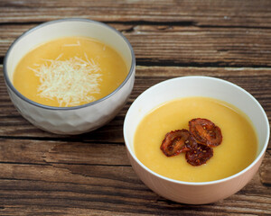 two plates of pumpkin cream soup with sprinkled cheese and pieces of dried tomato stand on a wooden table. diet lunch vitamins gluten free. side view