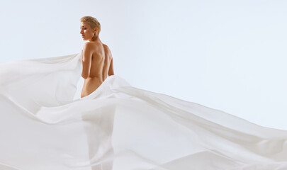 Back view portrait of young tender, fit woman body covering with white transparent floating cloth...