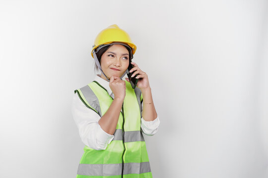 A thoughtful young woman labor worker wearing safety helmet and vest while getting a phone call and hand on her chin, isolated by white background. Labor's day concept.