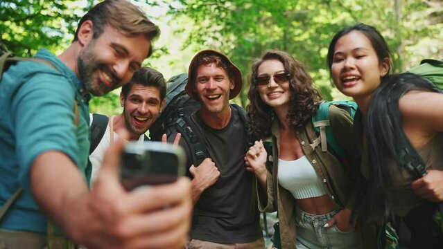 Smiling multinational backpacking friends taking selfies on cell phone near mountain river in forest