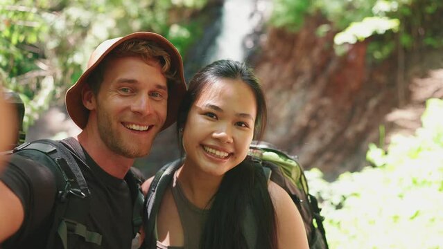 Smiling multinational tourist couple recording video on camera and greeting against waterfall in mountains