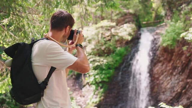 Back view of man tourist takes picture of waterfall on camera in mountains