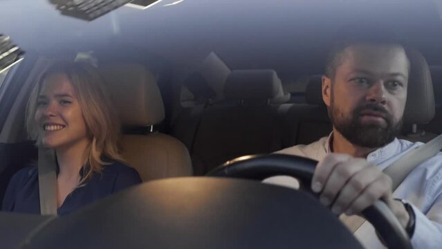 A young blonde woman and a handsome bearded man are driving in a luxury car with white leather seats and talking. Night view of the car interior through the windshield
