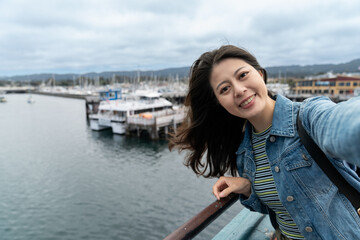Fototapeta na wymiar self portrait of happy asian woman visitor smiling at camera while taking selfie picture with boats near fishing port at Old Fisherman's Wharf in Monterey California usa