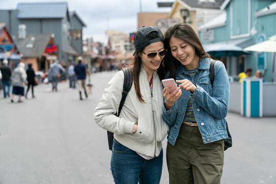 Two Smiling Asian Female Visitors Using Phone Together With Pointing Gesture On Street At Old Fisherman's Wharf In California Usa. They Are Having Pleasant Discussion About Their Travel Plan