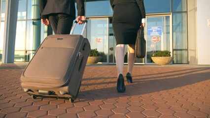 Business man and woman going to the airport with their luggage. Young businessman carrying suitcase on wheels and walking with his female colleague to terminal hall. Trip or travel concept Slow motion