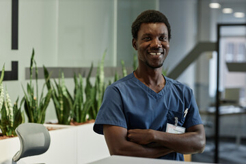 Young smiling African American clinics receptionist with crossed arms looking at camera while standing by counter in hall of modern hospital