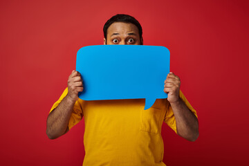 indian man with eyes wide open hiding behind blank speech bubble on red background, emotional