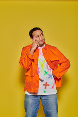 portrait of pensive indian man in orange jacket posing with hand on hip on yellow background