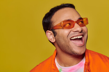 positive indian man in orange sunglasses looking away and smiling on yellow background, side glance