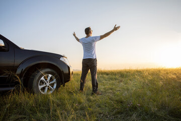 Happy man with arms up standing near his car and watching the sunset.  travel summer vacation. freedom concept.