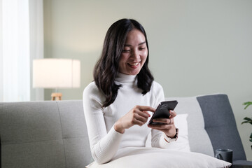 Fototapeta na wymiar Happy young asian woman relax on comfortable couch at home texting messaging on smartphone, smiling girl use cellphone, chatting online message.