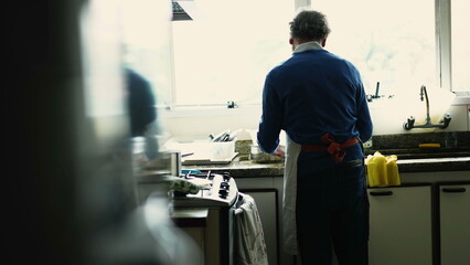 Back of senior man by kitchen sink preparing meal. Candid elderly cook at home cooking food by stove