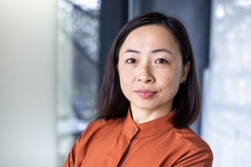 Close-up photo portrait of a young beautiful confident Asian woman standing in an office center and seriously looking at the camera.