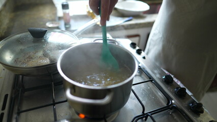 Close-up hand stirring food in boiling water inside metal pan at kitchen stove, meal preparation